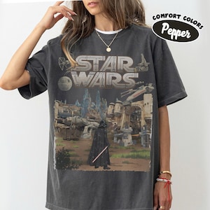 Può includere: T-shirt grigio scuro con grafica di Star Wars. Il design include il logo di Star Wars, Darth Vader con una spada laser e un paesaggio dettagliato. La maglietta è etichettata "Comfort Colors Pepper".