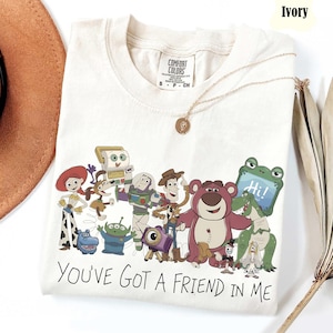 May include: An ivory short-sleeved t-shirt featuring a graphic print of various Toy Story characters including Woody, Buzz Lightyear, and Jessie. The text "YOU'VE GOT A FRIEND IN ME" is printed in black below the characters. The shirt has a crew neckline.