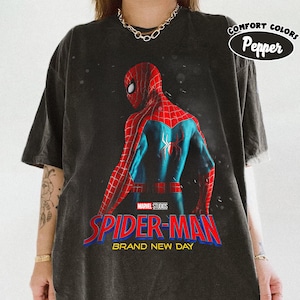 May include: Dark gray t-shirt featuring a graphic of Spider-Man in a red and blue suit, with the text "SPIDER-MAN BRAND NEW DAY" in red and blue. The shirt also has the text "COMFORT COLORS Pepper" in a white oval.