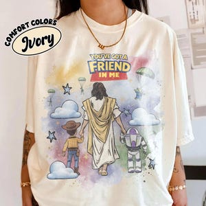May include: Ivory Comfort Colors t-shirt featuring a colorful graphic with the text "YOU'VE GOT A FRIEND IN ME." The design includes illustrations of Jesus, Woody, and Buzz Lightyear, with clouds, stars, and parachutes.
