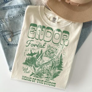 May include: Cream-coloured t-shirt with a green graphic featuring the text "ENDOR Forest" and an Ewok illustration. The shirt also includes the phrases "Yub Nub" and "HOME OF THE EWOKS". The shirt is folded and placed with a pair of jeans and a hat.