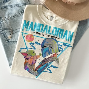 Może przedstawiać: Kremowy t-shirt z grafiką Mandalorian i Grogu, z napisem "MANDALORIAN" w kolorze turkusowym. Projekt zawiera tr&oacute;jkąt z r&oacute;żową i ż&oacute;łtą planetą. Koszulka jest złożona na niebieskich dżinsach, z naszyjnikiem i beżowym kapeluszem.
