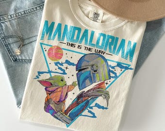 Op de afbeelding: Crèmekleurig T-shirt met een afbeelding van The Mandalorian en Grogu, met de tekst "MANDALORIAN" in turquoise. Het ontwerp bevat een driehoek met een roze en gele planeet. Het shirt is opgevouwen op een spijkerbroek, met een ketting en een beige hoed.