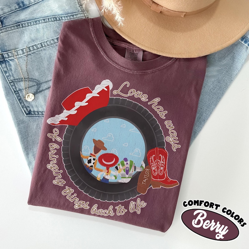 May include: A berry-colored t-shirt featuring a graphic of a tire framing a scene with three cartoon characters. The design includes a red cowboy hat, a red cowboy boot, and the text "Love has ways of bringing things back to life."