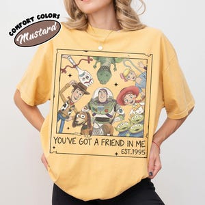 May include: Mustard-colored t-shirt featuring a framed graphic of Toy Story characters. The graphic includes Woody, Buzz Lightyear, and other characters with the text "YOU'VE GOT A FRIEND IN ME EST.1995". The shirt has short sleeves and a relaxed fit.