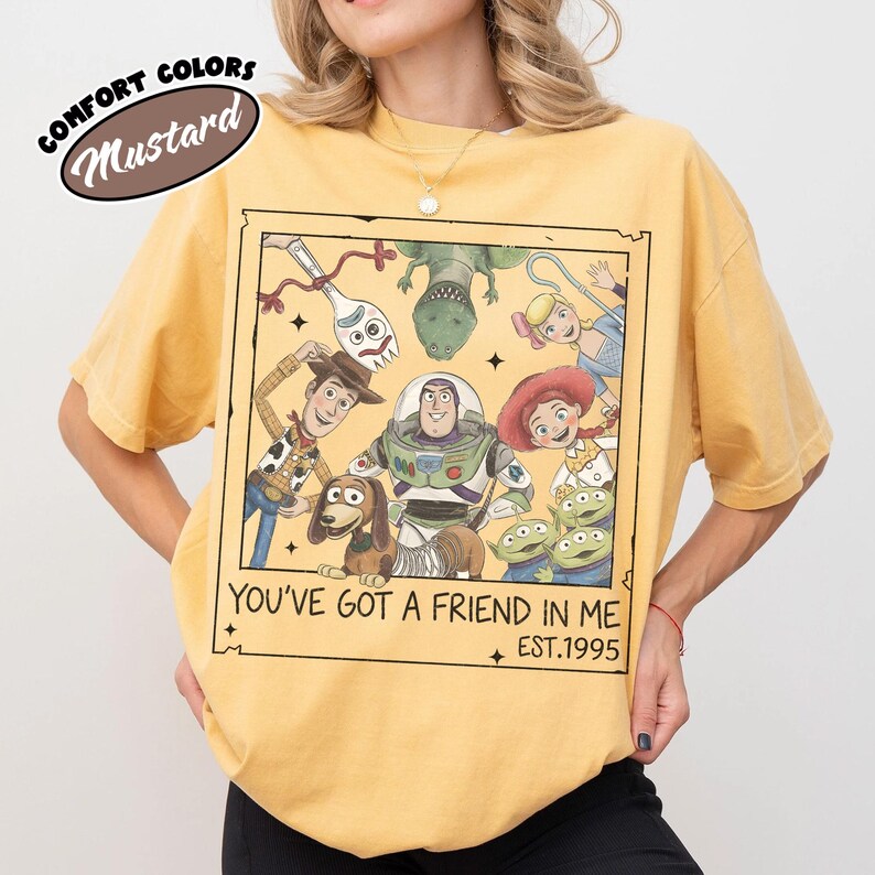 May include: Mustard-colored t-shirt featuring a framed graphic of Toy Story characters. The graphic includes Woody, Buzz Lightyear, and other characters with the text "YOU'VE GOT A FRIEND IN ME EST.1995". The shirt has short sleeves and a relaxed fit.