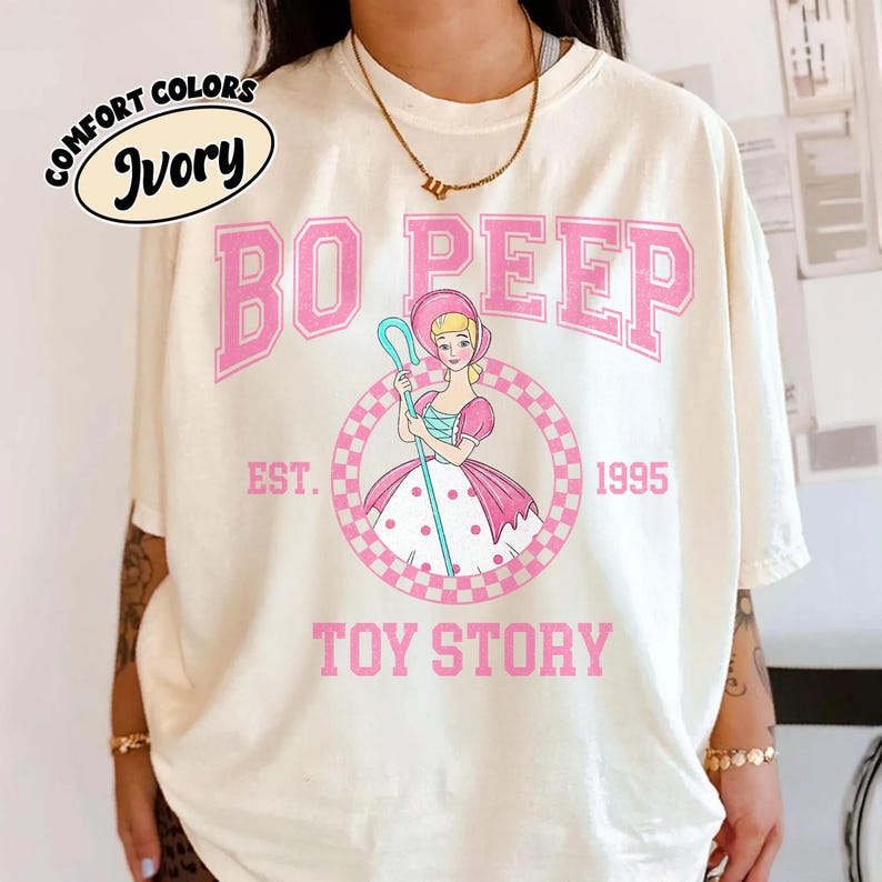 Pu&ograve; includere: T-shirt color avorio con grafica rosa di Bo Peep di Toy Story. Il design include il testo "BO PEEP", "EST. 1995" e "TOY STORY". La maglietta presenta anche le parole "COMFORT COLORS" e "Ivory" in un cerchio.
