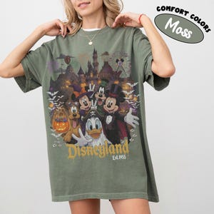May include: A moss green t-shirt featuring a graphic print of Disney characters in Halloween costumes, including Mickey Mouse, Minnie Mouse, Donald Duck, Goofy, and Pluto. The design includes a castle, fireworks, bats, and the text Disneyland Est. 1955.