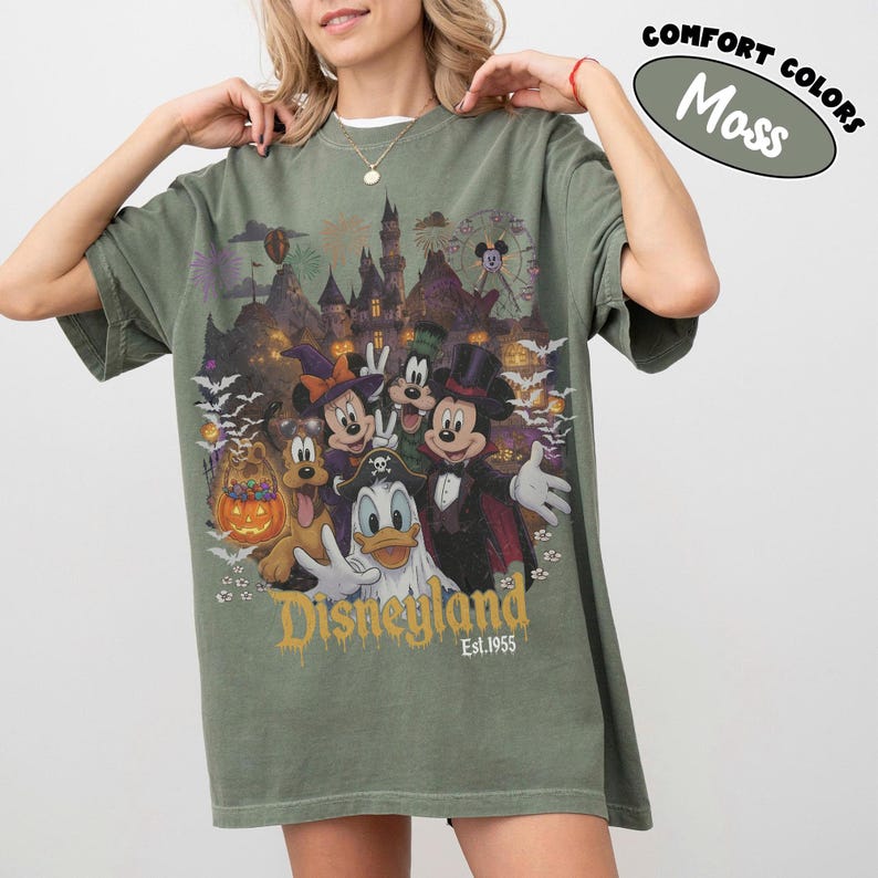 May include: A moss green t-shirt featuring a graphic print of Disney characters in Halloween costumes, including Mickey Mouse, Minnie Mouse, Donald Duck, Goofy, and Pluto. The design includes a castle, fireworks, bats, and the text Disneyland Est. 1955.