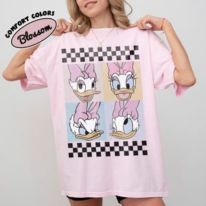 May include: A light pink t-shirt featuring a four-panel graphic of Daisy Duck with a black and white checkered pattern. The shirt has the words "COMFORT COLORS" and "Blossom" in a circular design.