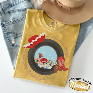 May include: A mustard yellow t-shirt featuring a graphic of a black tire framing a scene with a cowboy hat, a red boot, and cartoon characters. The text reads Love hat ways and bringing things back to life. The shirt has a Comfort Colors label.