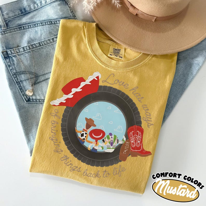 May include: A mustard yellow t-shirt featuring a graphic of a black tire framing a scene with a cowboy hat, a red boot, and cartoon characters. The text reads Love hat ways and bringing things back to life. The shirt has a Comfort Colors label.