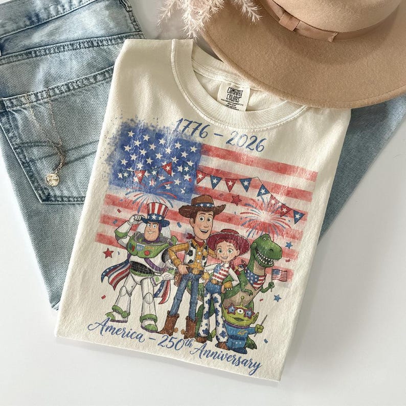 Może przedstawiać: Kremowa koszulka z grafiką amerykańskiej flagi i postaciami z filmu Toy Story. Koszulka ma napis "1776-2026" i "America - 250th Anniversary". Koszulka jest złożona na parze niebieskich dżins&oacute;w.