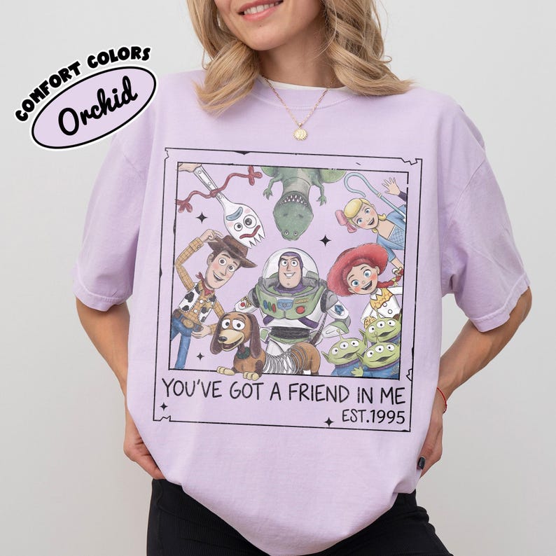May include: Orchid-colored t-shirt featuring a graphic of Toy Story characters within a framed design. The text "YOU'VE GOT A FRIEND IN ME" and "EST.1995" are included. The shirt is labeled "COMFORT COLORS".