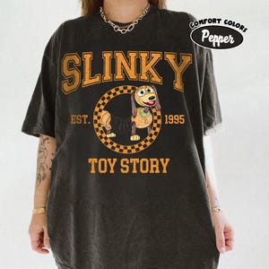 Pu&ograve; includere: T-shirt nero con un design arancione di Slinky Dog di Toy Story. Il design include la parola "SLINKY", il personaggio del cane all'interno di un cerchio a scacchi e il testo "TOY STORY EST. 1995". La maglietta ha un aspetto vintage.