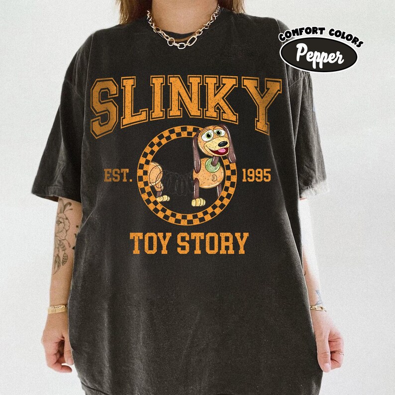 Pu&ograve; includere: T-shirt nero con un design arancione di Slinky Dog di Toy Story. Il design include la parola "SLINKY", il personaggio del cane all'interno di un cerchio a scacchi e il testo "TOY STORY EST. 1995". La maglietta ha un aspetto vintage.