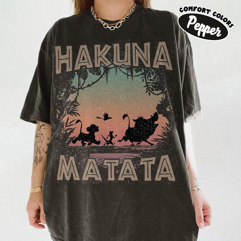 May include: Dark grey t-shirt with the words "HAKUNA MATATA" in a distressed font. The shirt features a colourful graphic of silhouettes of Simba, Timon, Pumbaa, and a bird. The brand name "Comfort Colors Pepper" is printed on the upper right.
