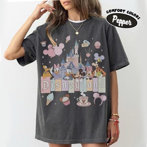 May include: A charcoal gray t-shirt featuring a graphic print of a castle, Mickey Mouse, Minnie Mouse, Donald Duck, Daisy Duck, Pluto, and Goofy. The text Disneyland is printed in the center. Additional icons include cupcakes, ice cream, and ears.