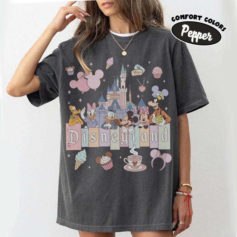 May include: A charcoal gray t-shirt featuring a graphic print of a castle, Mickey Mouse, Minnie Mouse, Donald Duck, Daisy Duck, Pluto, and Goofy. The text Disneyland is printed in the center. Additional icons include cupcakes, ice cream, and ears.
