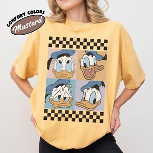 May include: Mustard-colored t-shirt featuring a four-panel graphic of Donald Duck's face with different expressions. The graphic is framed by a black and white checkered pattern. The text "COMFORT COLORS" and "Mustard" are in a circular design at the top left.