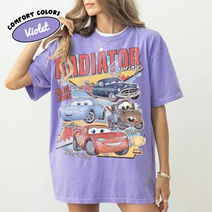 May include: A violet Comfort Colors t-shirt featuring a graphic of characters from the movie Cars. The design includes the text "Radiator Springs" and the names of the characters: Doc Hudson, Sally, Mater, and Lightning McQueen.