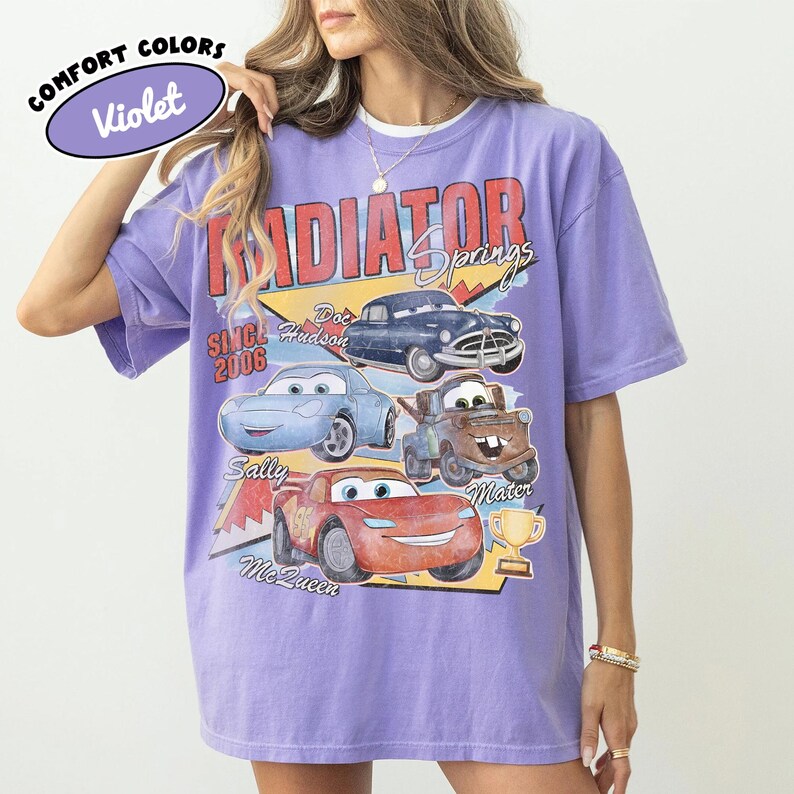 May include: A violet Comfort Colors t-shirt featuring a graphic of characters from the movie Cars. The design includes the text "Radiator Springs" and the names of the characters: Doc Hudson, Sally, Mater, and Lightning McQueen.