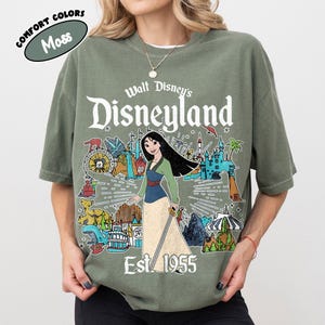 Op de afbeelding: Een mosgroen t-shirt met korte mouwen en een grafische print van Mulan met een zwaard, omringd door Disneyland-parkiconen en de tekst Walt Disney's Disneyland Est. 1955. Het shirt bevat een klein rond label met de tekst Comfort Colors Moss.