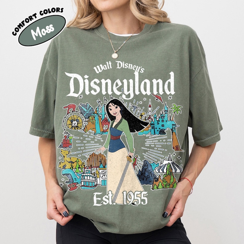 Op de afbeelding: Een mosgroen t-shirt met korte mouwen en een grafische print van Mulan met een zwaard, omringd door Disneyland-parkiconen en de tekst Walt Disney's Disneyland Est. 1955. Het shirt bevat een klein rond label met de tekst Comfort Colors Moss.