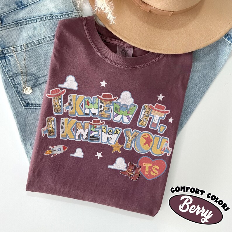 May include: A berry-colored t-shirt featuring a graphic print with the text "I KNEW IT, I KNEW YOU". The design includes illustrations of cowboy hats, a space ranger suit, a rocket, clouds, stars, cowboy boots, and a heart with the letters TS.