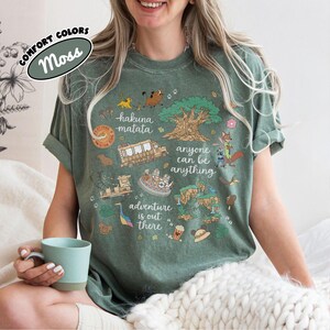 Puede incluir: Camiseta verde musgo con un dise&ntilde;o gr&aacute;fico colorido. El dise&ntilde;o incluye las palabras "hakuna matata", "anyone can be anything" y "adventure is out there", as&iacute; como varias ilustraciones de dibujos animados.
