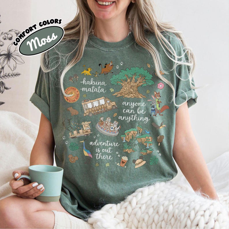 Puede incluir: Camiseta verde musgo con un dise&ntilde;o gr&aacute;fico colorido. El dise&ntilde;o incluye las palabras "hakuna matata", "anyone can be anything" y "adventure is out there", as&iacute; como varias ilustraciones de dibujos animados.