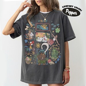 May include: Dark grey t-shirt with a colourful Disney character design. The shirt features the word "Disney" at the top and the words "Comfort Colors Pepper" in an oval. The design includes images of Moana, Maui, and other characters.