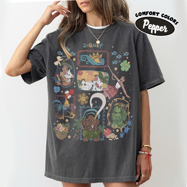 May include: Dark grey t-shirt with a colourful Disney character design. The shirt features the word "Disney" at the top and the words "Comfort Colors Pepper" in an oval. The design includes images of Moana, Maui, and other characters.
