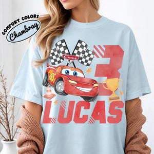 Puede incluir: Camiseta azul claro con un coche de carreras de dibujos animados rojo, el n&uacute;mero 95 y el nombre "LUCAS" en rojo. Tambi&eacute;n se muestra el n&uacute;mero "3" y un trofeo dorado. La camiseta tiene el texto "COMFORT COLORS Chambray".