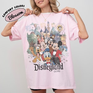 May include: A light pink t-shirt featuring a colorful graphic of Disney characters in front of a castle and a Ferris wheel. The word "Disneyland" is printed in large letters, with "Est.1955" below. The words "Comfort Colors" and "Blossom" are also printed on the shirt.