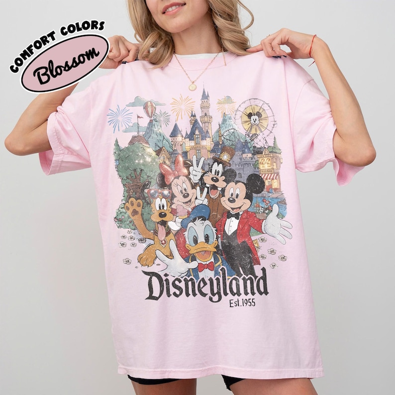 May include: A light pink t-shirt featuring a colorful graphic of Disney characters in front of a castle and a Ferris wheel. The word "Disneyland" is printed in large letters, with "Est.1955" below. The words "Comfort Colors" and "Blossom" are also printed on the shirt.
