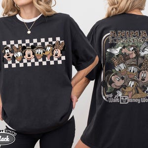 K&ouml;nnte beinhalten: Schwarzes T-Shirt mit einer Grafik von Disney-Figuren, darunter Mickey, Minnie, Donald, Goofy und Pluto, in Safari-Kleidung. Das Frontdesign ist vor einem schwarz-wei&szlig;en Schachbrettmuster platziert. Auf der R&uuml;ckseite des Shirts steht der Text "ANIMAL KINGDOM" und "Walt Disney World".