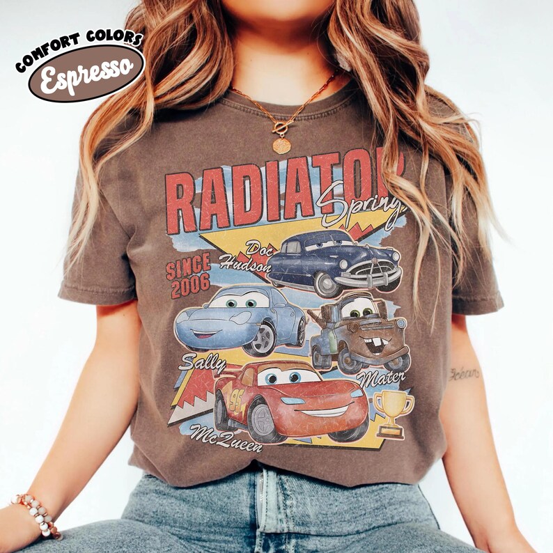May include: Espresso-colored t-shirt featuring a graphic of characters from the movie Cars. The design includes the text "RADIATOR SPRING" and the names of characters like Sally, McQueen, and Mater. The shirt also has the text "SINCE 2006".