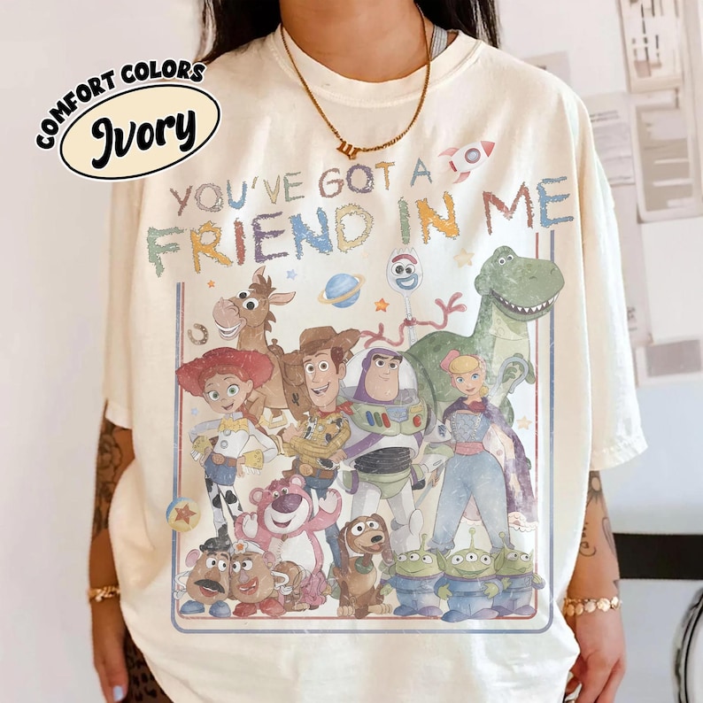 May include: An ivory t-shirt featuring a graphic print of various animated characters from the Toy Story franchise. The text "YOU'VE GOT A FRIEND IN ME" is printed above the characters in a multi-colored, distressed font style.