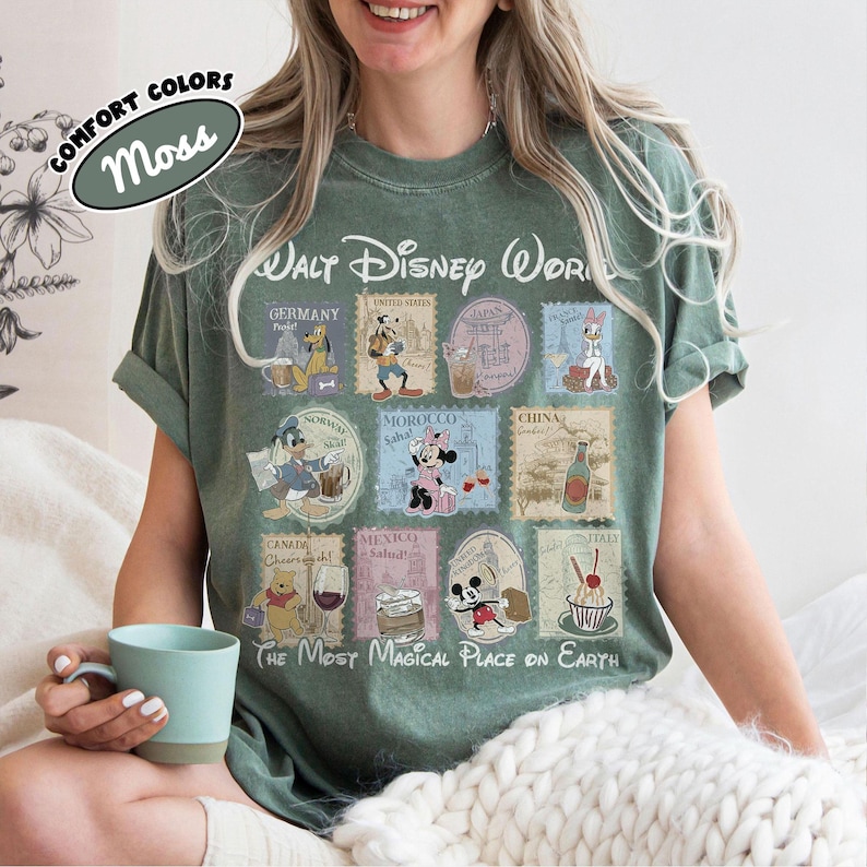 May include: Moss green t-shirt with a Walt Disney World design featuring stamps from various countries. The shirt includes the text "The Most Magical Place on Earth" and "Comfort Colors".