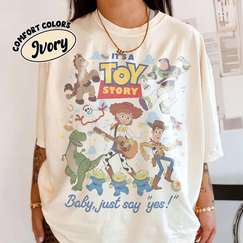 May include: An ivory t-shirt featuring a colorful graphic print of Toy Story characters including Woody, Jessie playing a guitar, Buzz Lightyear, Rex, Bullseye, Forky, and three aliens. The text reads "TOY STORY" and "Baby, just say yes!"