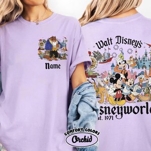 May include: A lilac Comfort Colors t-shirt featuring a Beauty and the Beast graphic on the front and a Disney World design on the back. The front has a Beauty and the Beast graphic with the word "Name" below. The back has Disney characters and the text "Walt Disney's Disneyworld est. 1971".