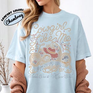 May include: A light blue t-shirt featuring a graphic print with the text "Cowgirl like Me" and "Takes One to Know One". The design includes rope borders, a cowboy hat, roses, a horseshoe, musical notes, and small stars in tan and red.