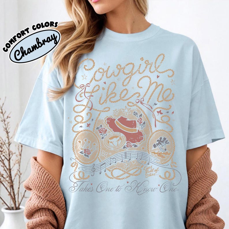May include: A light blue t-shirt featuring a graphic print with the text "Cowgirl like Me" and "Takes One to Know One". The design includes rope borders, a cowboy hat, roses, a horseshoe, musical notes, and small stars in tan and red.