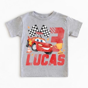 Puede incluir: Camiseta gris con un dise&ntilde;o de coche de carreras rojo, el n&uacute;mero 95, banderas a cuadros y un trofeo dorado. Tambi&eacute;n incluye el n&uacute;mero 3 y el nombre "LUCAS" en rojo. Un dise&ntilde;o divertido para el cumplea&ntilde;os de un ni&ntilde;o.