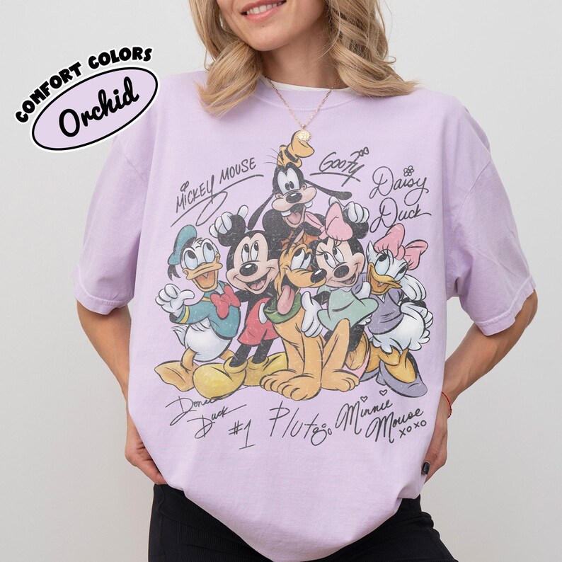 May include: A light purple t-shirt featuring a graphic of Mickey Mouse, Donald Duck, Goofy, Pluto, Minnie Mouse, and Daisy Duck. The shirt has the text "COMFORT COLORS Orchid" in a white oval. The characters' signatures are also printed on the shirt.