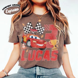 Puede incluir: Camiseta color espresso con un coche de carreras rojo de dibujos animados con el n&uacute;mero 95, banderas a cuadros y el nombre "LUCAS" en letras rojas. La camiseta tambi&eacute;n tiene un trofeo dorado y el n&uacute;mero 3.