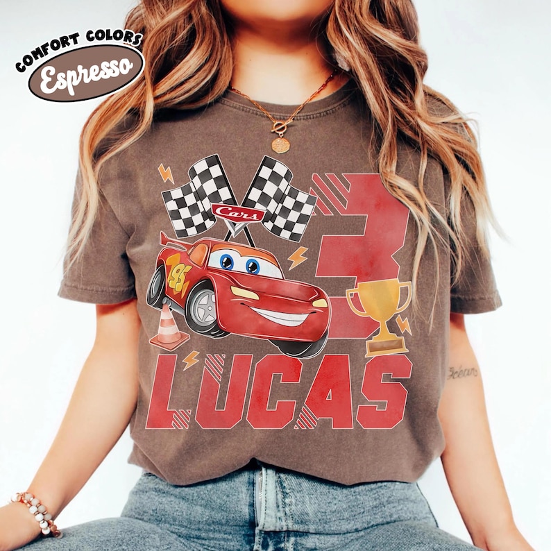 Puede incluir: Camiseta color espresso con un coche de carreras rojo de dibujos animados con el n&uacute;mero 95, banderas a cuadros y el nombre "LUCAS" en letras rojas. La camiseta tambi&eacute;n tiene un trofeo dorado y el n&uacute;mero 3.