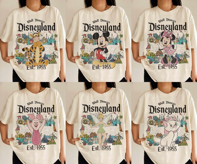 Op de afbeelding: Een cr&egrave;mekleurig t-shirt met een afbeelding van Walt Disney's Disneyland Est. 1955 met diverse personages zoals Teigetje, Mickey Mouse, Minnie Mouse, Knorretje, Tinkerbel en Marie, tegen een kleurrijke achtergrond van het park.