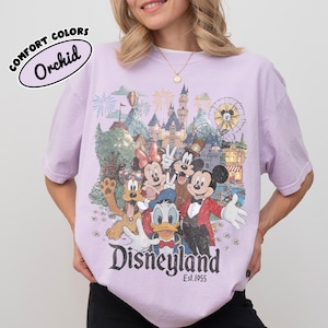 May include: Orchid Comfort Colors t-shirt featuring a Disneyland graphic with Mickey Mouse, Minnie Mouse, Donald Duck, Goofy, and other characters. The graphic includes a castle, fireworks, and the text "Disneyland Est. 1955."